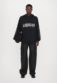 Hoodie negro de gran tamaño con texto gráfico blanco, impresiones laterales y un bolsillo delantero. Combinado con pantalones cargo negros y zapatos negros.