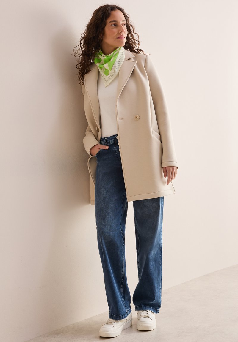 Beige skræddersyet frakke, hvid top, blå vide jeans, hvide sneakers og et grønt mønstret tørklæde, poseret foran en lys væg.