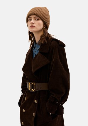 Cappotto trench in corduroy marrone con ampio colletto, bottoni grandi e cintura coordinata. Abbinato a un berretto a coste marrone e una camicia di jeans.