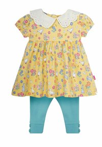 Keltainen mekko, jossa on kukkakuosi ja valkoinen pitsikaulus, yhdistettynä sinisiin ribattuihin legginsseihin. Leggien hihoissa on nappikoristeita.