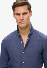 Marineblauwe linnen shirt met een staande kraag, subtiele textuur, knoopsluiting aan de voorkant en een klein geborduurd logo op de linkerborst.