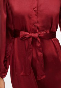 Bordowa sukienka z satyny z zapięciem na guziki z przodu, długimi rękawami i paskiem wiązanym w talii, o gładkiej fakturze i delikatnym połysku.