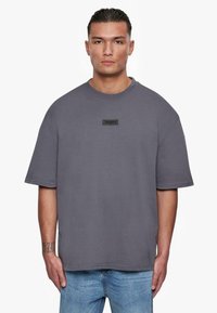Grå oversize t-shirt med korte ærmer, der har et sort rektangulært logo-patch på brystet; lavet af glat stof med et afslappet fit.