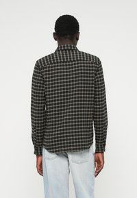 Άνδρας με κοντά σγουρά μαλλιά που φοράει πουκάμισο με μαύρες και μπεζ καρό λεπτομέρειες και ανοιχτό μπλε τζιν, στέκεται με την πλάτη γυρισμένη σε λευκό φόντο.