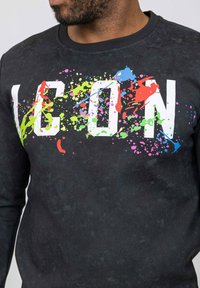 Sweatshirt noir avec un texte "ICON" blanc en gras, éclaboussé de couleurs vives comme le rose, le vert et le bleu, doté d'une texture douce et d'un col côtelé.