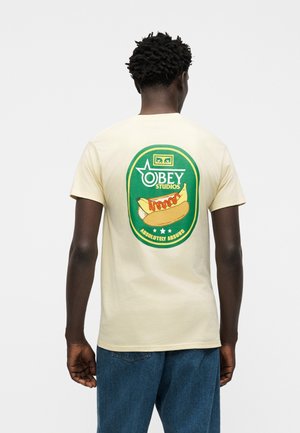 Sötét bőrű, rövid, göndör hajú személy, bézs pólót visel, amelyen zöld és sárga hot dog grafika és az "Obey Studios Absolutely Absurd" felirat található a háton.