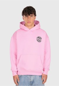 Junger Mann mit lockigem Haar trägt einen rosa Hoodie mit einer Fronttasche und schwarzem "Drop Size"-Text auf der linken Brust, kombiniert mit hellblauen Jeans.