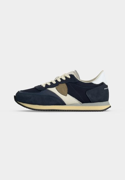 Baskets bleu marine et blanches avec lacets beiges, empiècements en daim et en mesh, semelle plate et design rétro de chaussure de course montrées de profil.