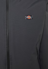 Veste noire zippée en tissu texturé, avec une petite étiquette de marque sur la poitrine et une poche latérale à ouverture diagonale.