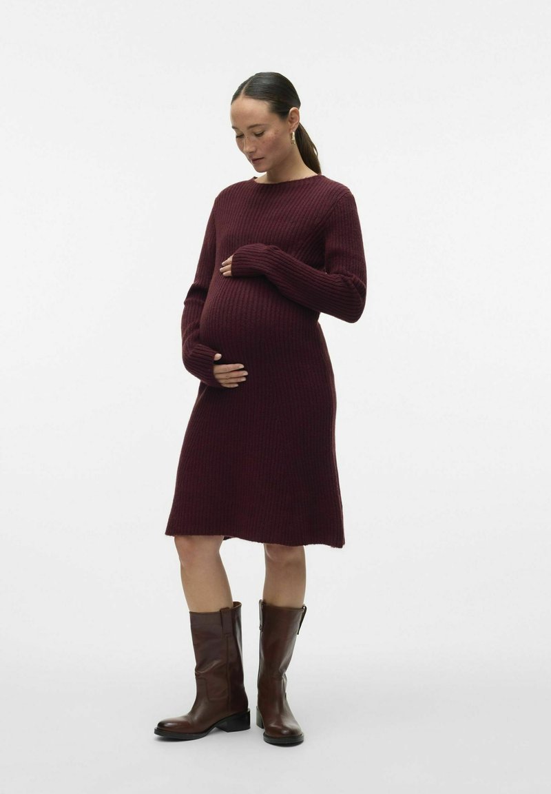 Vestito in maglia a coste per maternità di colore bordeaux, con maniche lunghe e orlo leggermente svasato. Abbinato a stivali al ginocchio in pelle marrone.