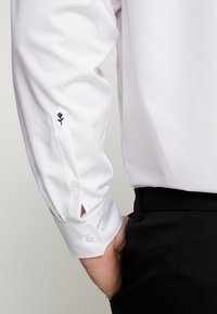 Witte langemouwen shirt met knoopmanchetten, voorzien van een geborduurd zwart rozenontwerp nabij de manchetten. Gladde stof, formele stijl.