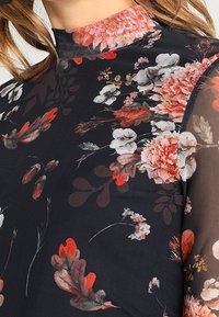 Blumenbluse mit hohem Kragen, transparenter Stoff und schwarzem Hintergrund. Verziert mit rosa, weißen und grünen Blumen in einem gemischten Muster.