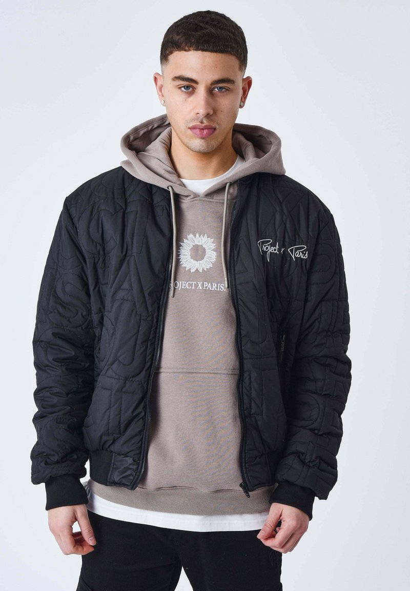 Jeune homme de face portant une veste matelassée noire sur un hoodie taupe avec un tournesol et le logo "Project X Paris", les mains détendues le long du corps.