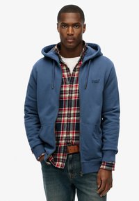 Sudadera azul con cordones sobre una camisa de franela de cuadros rojos y azules. La sudadera cuenta con una cremallera y bolsillos frontales. Unos pantalones vaqueros completan el look.