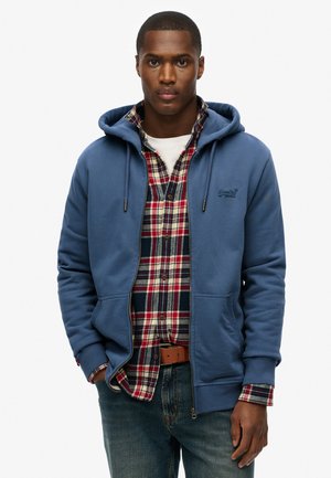 Felpa blu con cordini sopra una camicia di flanella a quadri rossi e blu. La felpa presenta una zip e tasche frontali. I jeans in denim completano il look.