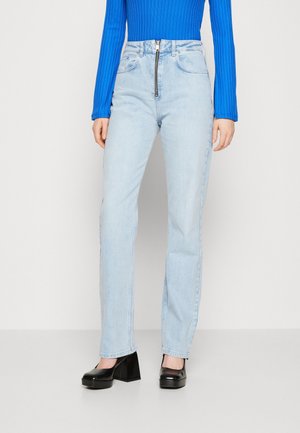 Kobieta ubrana w jasnoniebieskie dżinsy z wysokim stanem i przednim zamkiem błyskawicznym, niebieski prążkowany top z długim rękawem oraz czarne buty na platformie z obcasem.