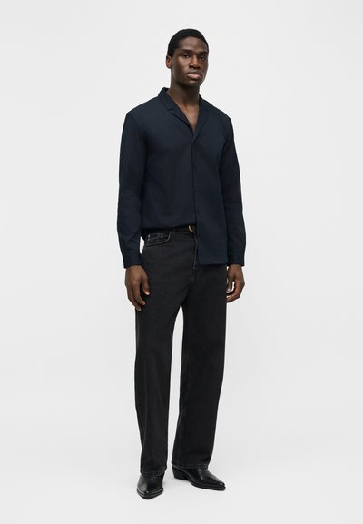 Mann trägt ein dunkelblaues, langärmeliges Hemd mit Knöpfen, schwarze Jeans und schwarze spitze Schuhe, steht vor einem schlichten grauen Hintergrund.