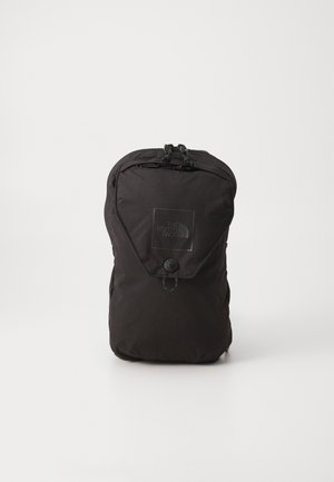 Sac à dos compact noir avec une poche avant à rabat et une fermeture éclair, avec le logo The North Face sur un fond blanc uni.
