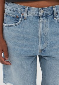 Gros plan d'une personne portant un jean en denim bleu clair avec une taille boutonnée et des poches avant visibles, la main gauche reposant à ses côtés.