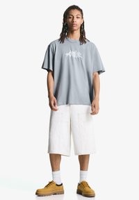 Grå oversized t-shirt med hvid grafik, parret med hvide knælange cargo shorts og brune snørebåndssko. Modellen står frontvendt.