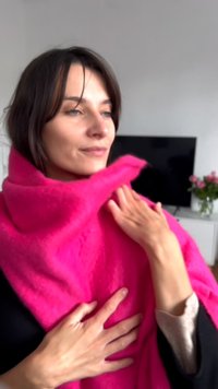 Puhaszőrű rózsaszín sál a vállon, lágy textúrával. A kezek a szövetet érintik, semleges háttér előtt.