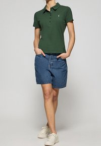 Zielona koszulka polo z kołnierzykiem i trzema guzikami, zestawiona z niebieskimi szortami dżinsowymi. Białe sneakersy dopełniają całość.