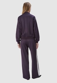 Tweedelig trainingspak van donkerpaars fabric. De jas heeft een hoge kraag; de broek heeft een witte zijkantstreep. Relaxte pasvorm, zachte textuur.