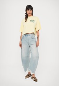 T-shirt jaune court avec texte vert, associé à un jean large déchiré bleu clair et des chaussures slip-on à imprimé léopard.