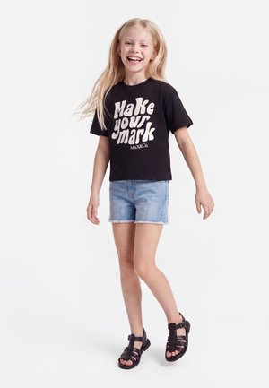 Uśmiechnięta dziewczyna z długimi blond włosami, ubrana w czarną koszulkę "Make your mark", jasnoniebieskie denimowe szorty i czarne sandały, stojąca na białym tle.