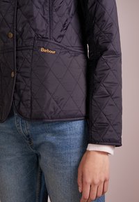 Marine gekwilt jack met ruitpatroon, drukknopen en Barbour-logo. Heeft een getailleerde uitstraling en zichtbare zakdetails.