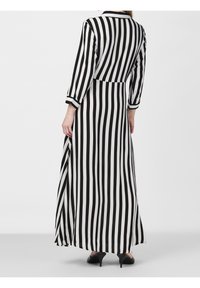 Femme portant une robe longue à rayures verticales noires et blanches avec manches longues et talons noirs, debout devant un fond uni.