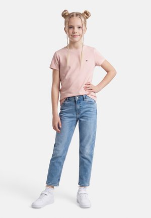 Młoda dziewczyna o blond włosach upiętych w dwa koki i warkocze, ubrana w różową koszulkę z krótkim rękawem, niebieskie dżinsy i białe trampki, stojąca z ręką na biodrze.