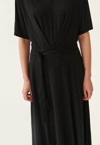 Schwarzes Wickelkleid aus weichem Stoff, mit V-Ausschnitt und kurzen Ärmeln. Es hat einen gebundenen Gürtel in der Taille und einen fließenden Rock.
