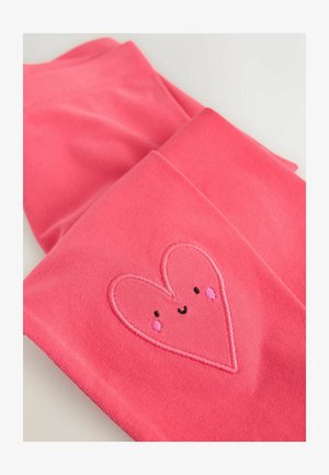 Colanți roz din material moale, cu un design brodat în formă de inimă, având obraji înroșiți și o față zâmbitoare.