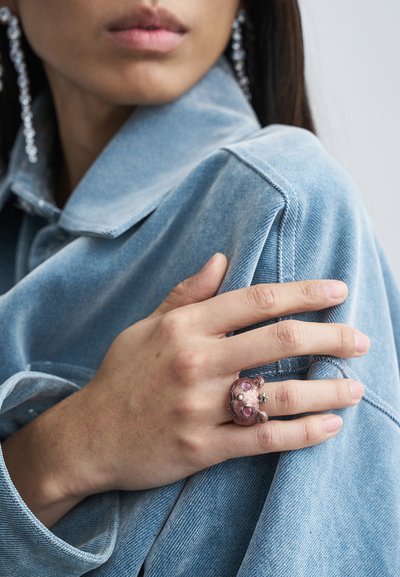 Close-up of a woman wearing a blue denim jacket and a pink bear-shaped ring with a small star on her finger, hand resting on her shoulder.
