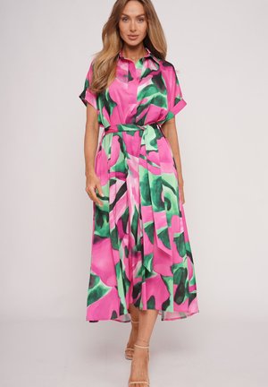 Woman with long hair wearing a belted pink dress with green and black abstract floral patterns and beige ankle-strap heels standing on a plain background.