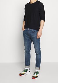Svart långärmad skjorta, blå slim-fit jeans och flerfärgade sneakers med vita sulor, gröna och gula detaljer, samt en vikbar manschett.