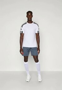 Tenue de sport Adidas comprenant un t-shirt blanc à manches courtes avec des rayures noires et un short gris. Le mannequin porte des chaussettes blanches et des chaussures de football.