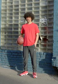 Camiseta atlética vermelha com logótipo, calças de jogging pretas e sapatilhas de basquetebol vermelhas. A pessoa segura uma bola de basquetebol em pé à frente de uma parede de bloco de vidro.