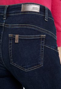 Los jeans de mezclilla de lavado oscuro presentan un pequeño acento metálico en el bolsillo trasero y un parche con el logo cosido en la cinturilla.