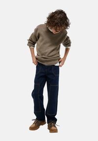 Un pull en tricot marron, un jean bleu foncé avec de grandes poches et des chaussures de randonnée beiges. L'ensemble présente une coupe décontractée et des matériaux texturés.