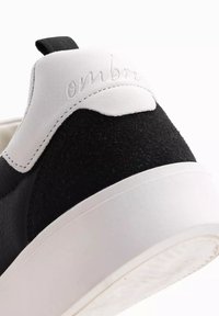 Svart lädersneaker med en texturerad svart mockapanel på baksidan och vit gummisul, med det präglade ordet "ombre" på hälen.