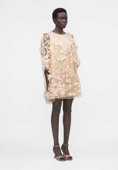 Γυναίκα στέκεται φορώντας μια διάφανη γυμνή φορεσιά με 3D λουλουδένιες εφαρμογές, συνδυασμένη με γυμνά πέδιλα με λουράκι στον αστράγαλο, μπροστά σε απλό λευκό φόντο.