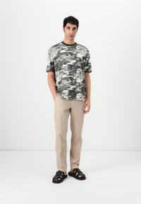 Grå camo T-skjorte med korte ermer, kombinert med lysebeige bukser og svarte sandaler med snøring og tykke såler.