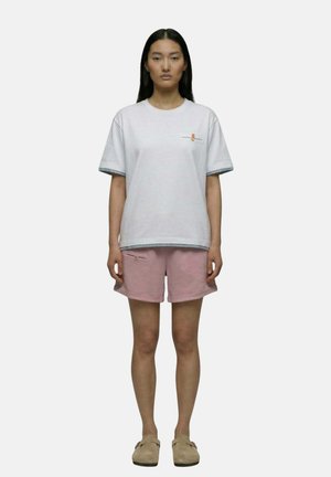 Femme debout face à l'avant, portant un t-shirt blanc ample, un short rose avec une poche zippée et des chaussures beige à enfiler, sur un fond uni.