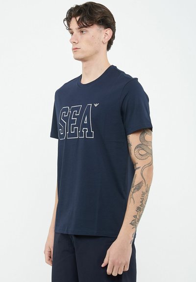 Giovane uomo con capelli scuri che indossa una maglietta blu navy con la scritta "SEA" e pantaloni scuri, mostra tatuaggi di serpenti e fiori sul braccio destro.