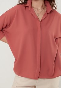 Frau in einer lockeren, kurzärmeligen Bluse in staubrosa mit einer mehrschichtigen Halskette und beigen Hosen vor einem schlichten Hintergrund.