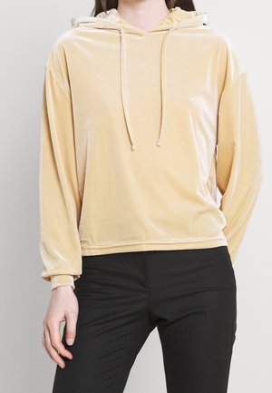 Femme portant un hoodie en velours jaune clair à manches longues avec cordons, associé à un pantalon noir ajusté sur un fond uni.