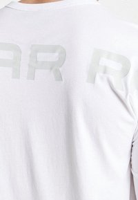 Vit t-shirt i mjukt tyg med matt yta. På baksidan finns stora, ljusgrå bokstäver i en fet, modern stil.