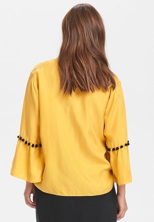 Kobieta z brązowymi włosami do ramion, ubrana w żółtą bluzkę z czarnym pomponowym wykończeniem na szerokich rękawach do łokcia, widziana od tyłu.